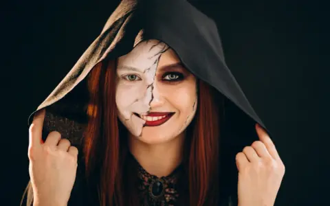 Kobieta w kapturze z połową twarzy pomalowaną na biało, z pęknięciami, druga połowa z ciemnym makijażem. Idealny prosty makijaż na Halloween.