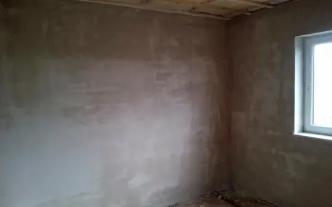 Unverputzte Innenwand mit Fensterecke und sichtbaren Deckenbalken.