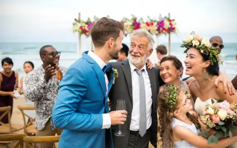 Szczęśliwy ojciec pana młodego w eleganckim garniturze śmieje się z synem na plażowym weselu.