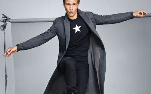 Joven con abrigo largo gris, camiseta negra con estrella blanca y vaqueros oscuros. Explora diversos tipos de abrigos para tu estilo.