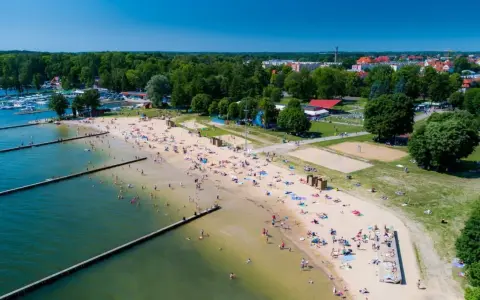 Słoneczna plaża w Giżycku tętni życiem. Ludzie odpoczywają na piasku, kąpią się w jeziorze i spacerują wzdłuż drewnianych pomostów.