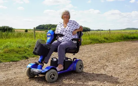 Uśmiechnięta kobieta na niebieskim skuterze elektrycznym inwalidzkim, ciesząca się dniem na wsi.