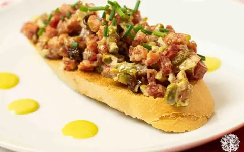 Tosta con steak tartar de salchichón y cebollino picado, decorada con puntos de salsa amarilla.