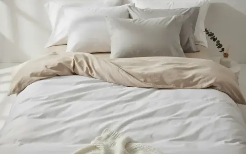 Ein Bett mit weißer und sandfarbener Bettwäsche aus Satin-Baumwolle. Mehrere Kissen liegen auf dem Bett.