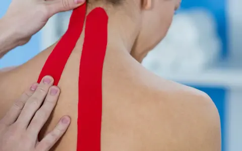 Czerwone plastry kinesio taping na karku i ramieniu, aplikowane przez fizjoterapeutę.