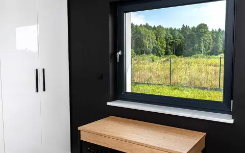 Biała szafa, czarna ściana i drewniane biurko pod oknem z białym parapetem. Za oknem zielony krajobraz.
