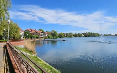 Spokojne jezioro zamkowe z fontanną i drewnianym pomostem, otoczone zielenią i zabudowaniami.