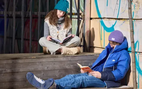 Dwoje dzieci czyta na schodkach. Dziewczynka w turkusowej czapce pochyla się nad książką, a chłopiec w fioletowej czapce przegląda lektury obowiązkowe.