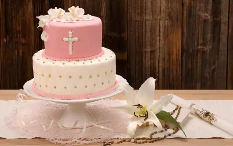 Elegancki dwupoziomowy tort na chrzest w różowo-białych barwach, ozdobiony krzyżem i kwiatami. Obok lilia, różaniec i świeca.