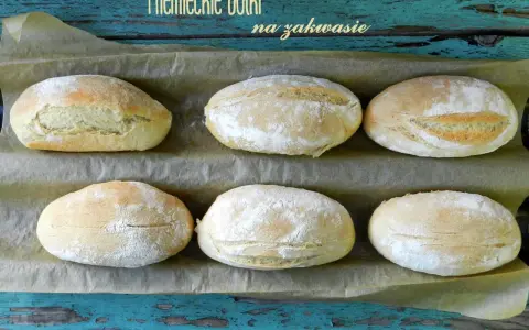 Sześć puszystych bułek na zakwasie, lekko oprószonych mąką, ułożonych na papierze do pieczenia.