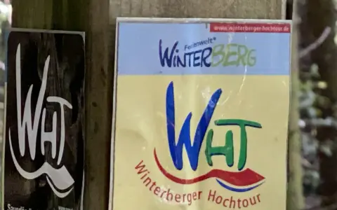 Schilder für die Winterberger Hochtour. Ein Schild zeigt "WHT" in Weiß auf Schwarz, das andere "Winterberg" und "Winterberger Hochtour" in bunten Farben.