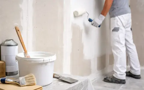 Un peintre applique une couche de peinture sur un mur en Fermacell. Matériel de peinture à proximité.