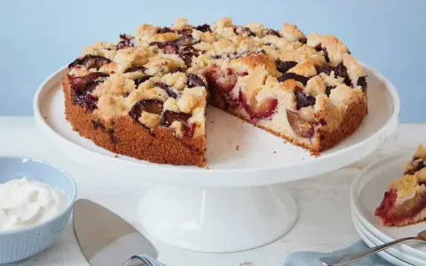 Ein einfacher Pflaumenkuchen mit Streuseln auf einem Tortenständer, daneben eine Schüssel Schlagsahne und ein Tortenheber.