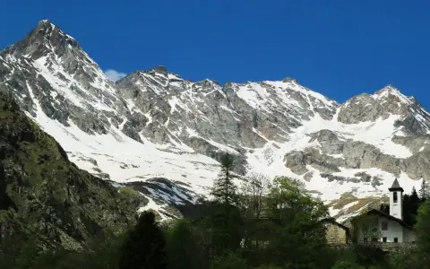 Imponente montagna sacra innevata, con un piccolo campanile bianco che emerge tra gli alberi verdi in primo piano.