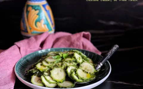 Zucchine marinate cotte, condite con erbe aromatiche, servite su un piatto verde. Sullo sfondo, un oggetto decorativo colorato e un tovagliolo rosa.