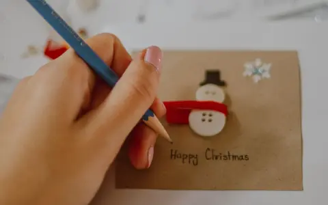 Ręka z ołówkiem pisze "Happy Christmas" na kartce z bałwankiem z guzików. Idealny przykład, jak narysować kartkę świąteczną.
