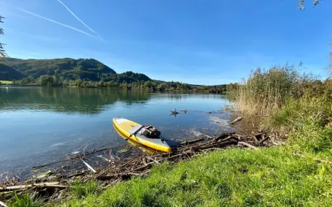 Gelbes SUP-Board liegt am Ufer bereit für SUP Touren Bayern. Ruhiger See, bewaldete Hügel und blauer Himmel.