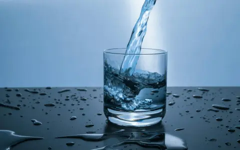Un jet d'eau de source calcaire tombe dans un verre, créant des éclaboussures. Des gouttes d'eau parsèment la surface sombre.
