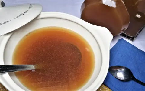 Caldo de carne casero en una cazuela blanca con tapa, junto a una jarra y cucharas.