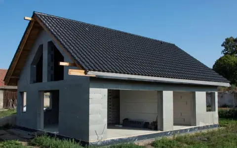 Dom w stanie surowym otwartym, z dachem pokrytym czarną dachówką. Ściany z bloczków betonowych, widoczne otwory okienne i drzwiowe.