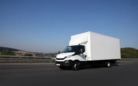 Biały samochód ciężarowy powyżej 3,5 tony pędzi autostradą. W tle krajobraz z pagórkami i domami.
