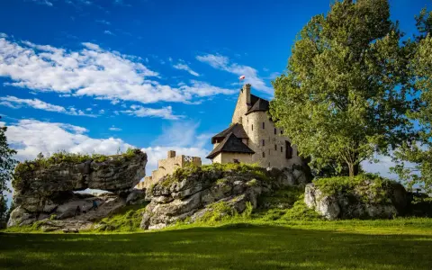Zamek Bobolice na skale, z polską flagą, pod błękitnym niebem z chmurami. Obok skalny most i zielona trawa.
