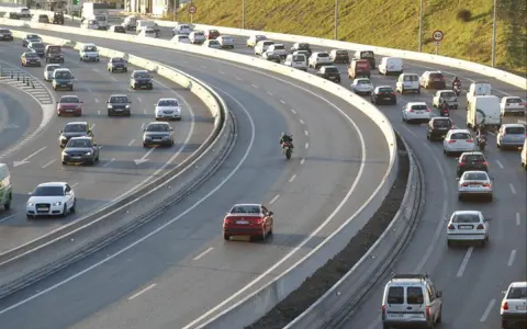 Tráfico intenso en una autopista con varios carriles. Coches, furgonetas y una moto forman la **circulación vao** en un día soleado.