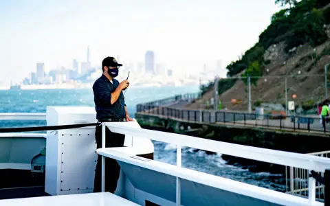 Un homme sur un bateau utilise une radio. Il est en communication, peut-être sur une fréquence vhf libre, avec la ville en arrière-plan.