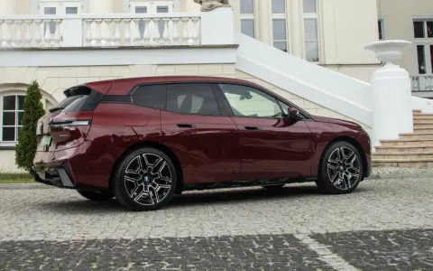 Ciemnoczerwone BMW iX stoi na brukowanym podjeździe przed eleganckim budynkiem.
