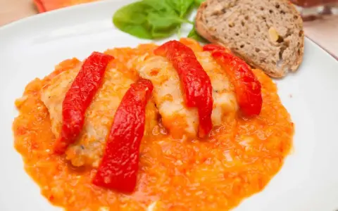 Bacalao a la riojana, un plato tradicional con trozos de pescado blanco cubiertos de pimientos rojos y una salsa de tomate y pimientos. Servido con pan y hojas verdes.