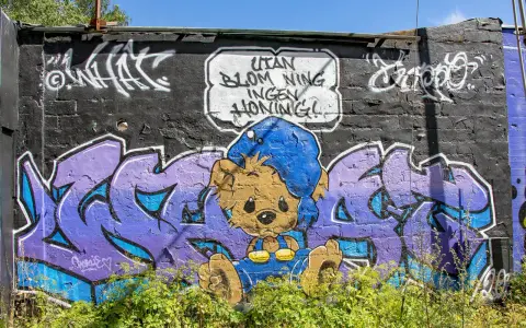 Enkel graffiti på en tegelvägg visar en brun nallebjörn i blå overall och mössa, med texten "Utan blommor ingen honung!".