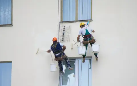 Deux peintres en bâtiment travaillent en hauteur sur une façade. L'un utilise un rouleau pour la peinture immeuble, l'autre est prêt à continuer.