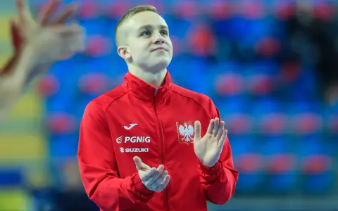Adam Jędraszek w czerwonej kurtce z orłem, z dłońmi uniesionymi w geście radości lub powitania.