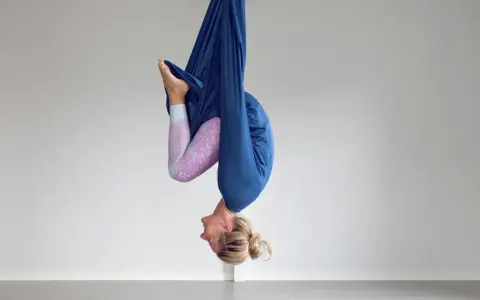 Kvinna hänger upp och ner i en blå tyghängmatta, utövar flying yoga.