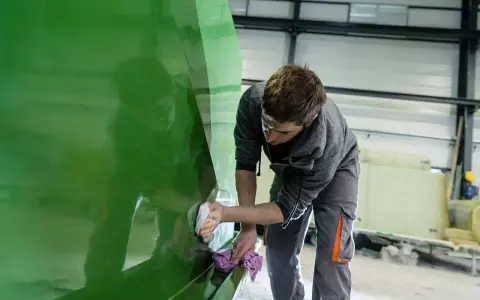 Un ouvrier polit la coque d'un bateau en polyester vert vif dans un atelier.