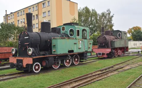 Dwa zabytkowe parowozy, zielony i rudy, stoją na torach przy budynku. To część ekspozycji muzeum kolei wąskotorowej w Sochaczewie.