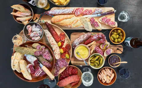 Eine reichhaltige Wurstplatte anrichten mit Baguette, Oliven, Cornichons und Senf.
