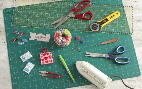 Matériel de couture : ciseaux, cutter rotatif, aiguilles, épingles, fils, fer à repasser, sur un tapis de découpe.