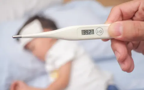 Cyfrowy termometr pokazuje 39.2°C, wskazując wysoką temperaturę ciała u chorego dziecka leżącego w łóżku.