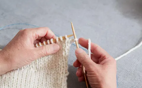 Händer stickar en vit resår med trästickor. En blå kabel syns i bakgrunden.