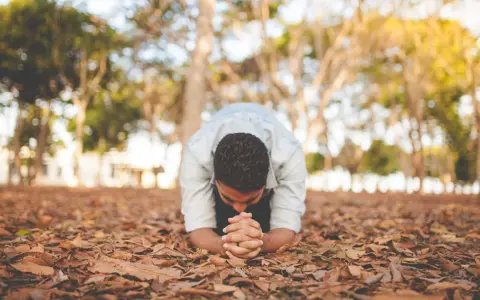 A man in a light blue shirt is in a posture of christian prostration, hands clasped on the ground covered in fallen leaves.