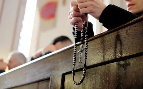 Dłonie trzymające różaniec podczas nabożeństwa. W tle widać innych wiernych, przygotowujących się na odpust.