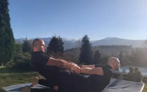 Dwóch mężczyzn wykonuje tajski masaż na macie na zewnątrz. W tle góry i las.