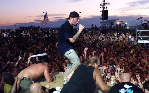 Tłum na Woodstock 99, z wokalistą na scenie i amerykańską flagą. Atmosfera była napięta, co doprowadziło do późniejszych zamieszek i ofiar.