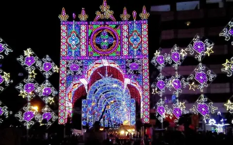 Un tunnel di luci colorate e decorazioni luminose, tipiche delle feste patronali, crea un'atmosfera magica e festosa.