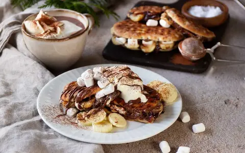 En härlig nutella macka med banan och marshmallows, toppad med grädde och kakao. Bredvid står en kopp varm choklad.