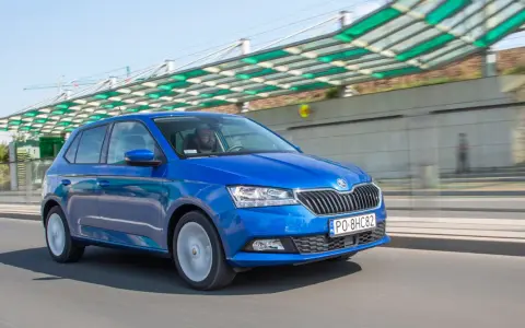 Niebieska Škoda Fabia jedzie po drodze, za nią widać nowoczesną konstrukcję z zielonym zadaszeniem.