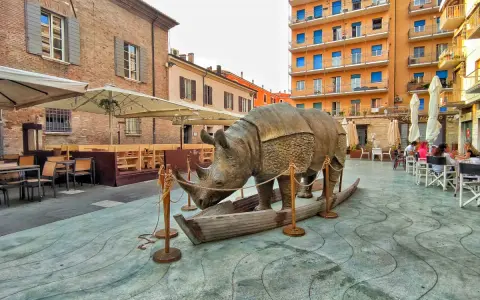 Statua di rinoceronte in una barca, in una piazza di Rimini, dove è nato Fellini. Tavolini all'aperto e palazzi colorati sullo sfondo.