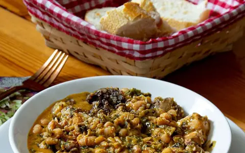 Un reconfortante cocido montañés, con garbanzos, verduras y trozos de carne, servido en un cuenco blanco. A su lado, una cesta con pan y un tenedor.