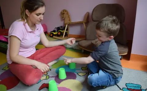 Terapia behawioralna autyzm: chłopiec i terapeutka bawią się kubeczkami na kolorowym dywanie.